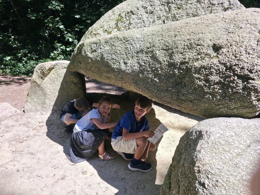 Drie kinderen gehurkt onder de zware dekstenen van het hunebed, lachend naar de camera