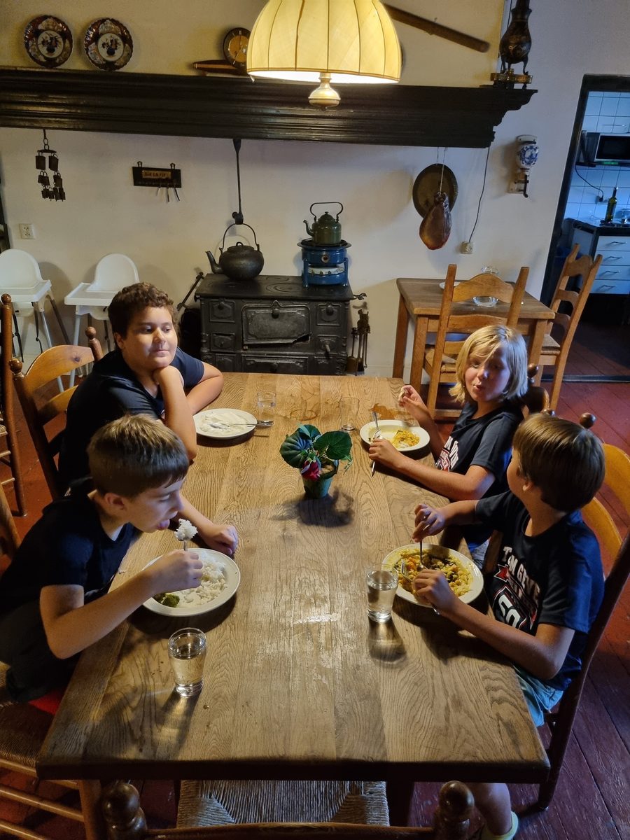 Vier jongens aan de lange houten tafel eten rijst met curry, onder de gele hanglamp met de oude houtkachel op de achtergrond