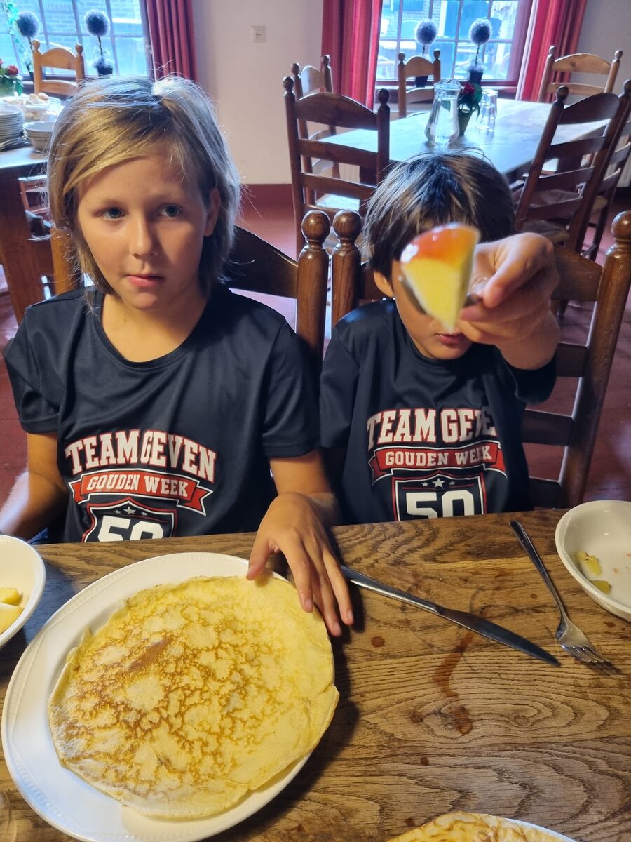 Twee jongens in donkerblauwe Team Geven Gouden Week-shirts aan de ontbijttafel met pannenkoeken, de voorste houdt een stukje fruit omhoog