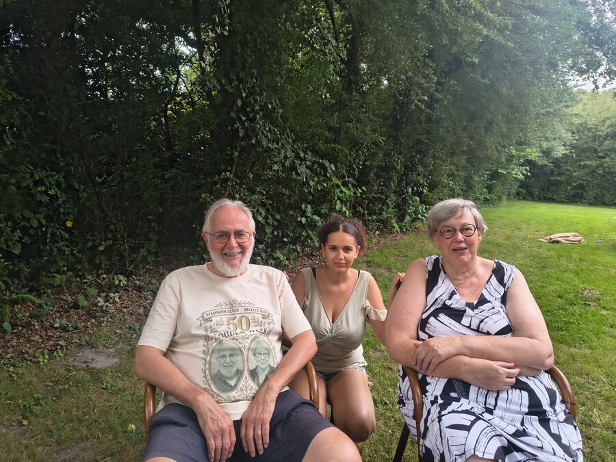 Een oudere man en vrouw zitten met een jonge vrouw ertussen op houten stoelen buiten, voor een groene haag