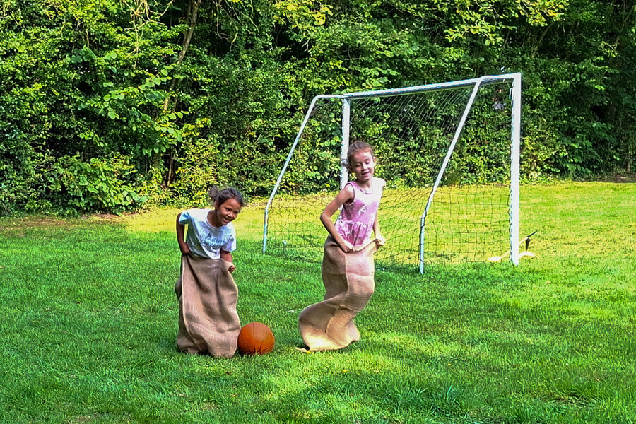 Twee meisjes springen lachend in jute zakken over het grasveld, met een voetbaldoel op de achtergrond