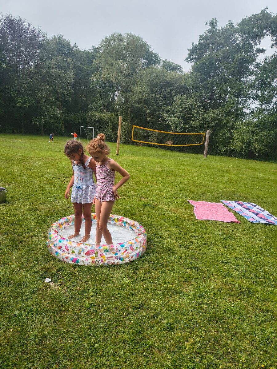 Twee meisjes staan in een klein opblaasbadje op het gazon, met een voetbaldoel en volleybalnet op de achtergrond