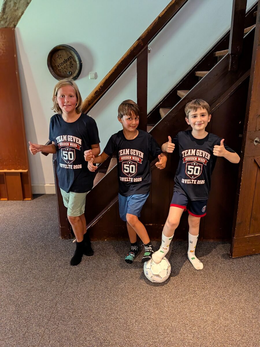 Drie jongens in het donkerblauwe Team Geven-shirt onder de trap, duimen omhoog, een voetbal onder een voet