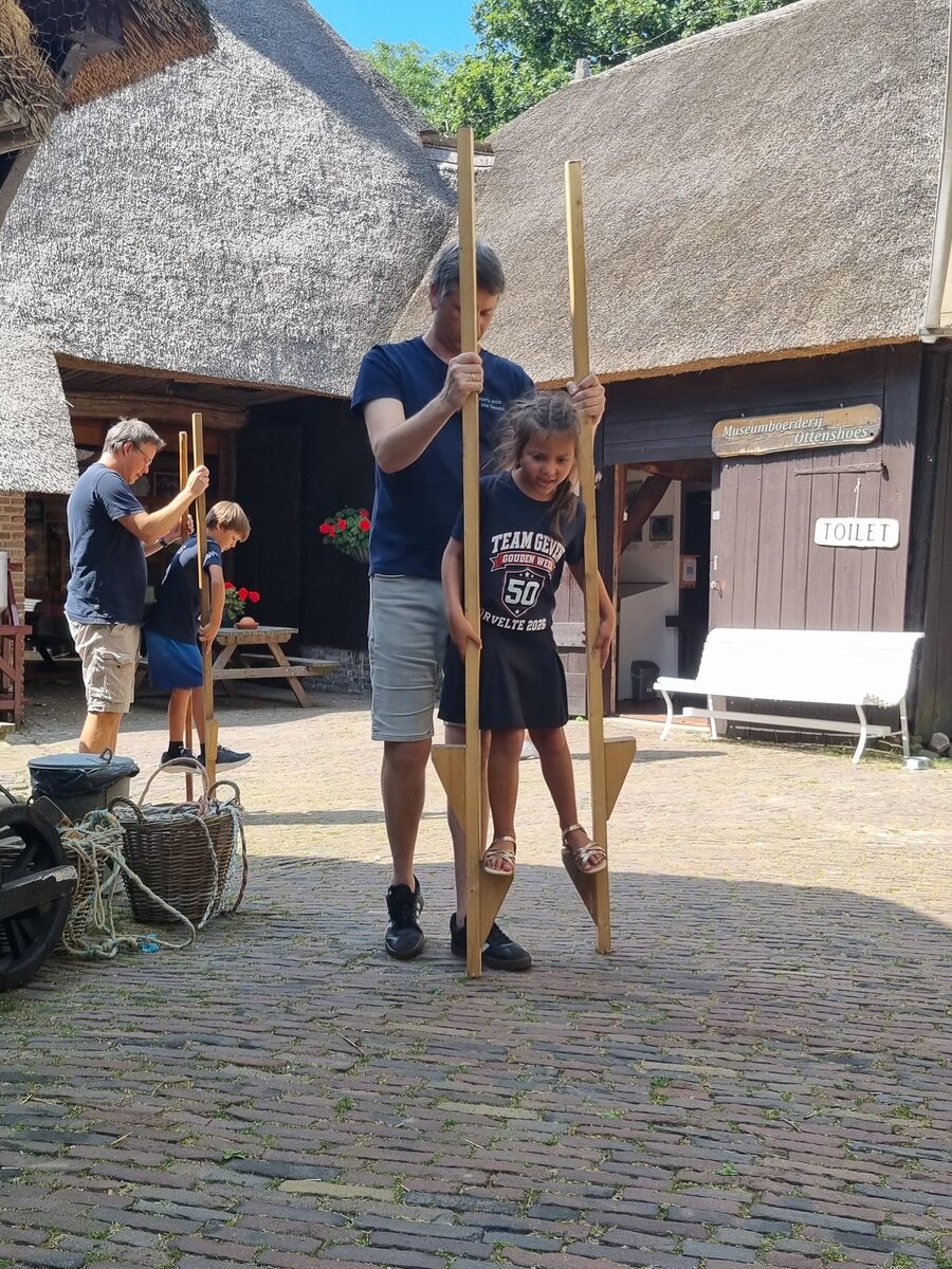 Arjan houdt Iris in evenwicht op houten stelten op de keien bij de museumboerderij