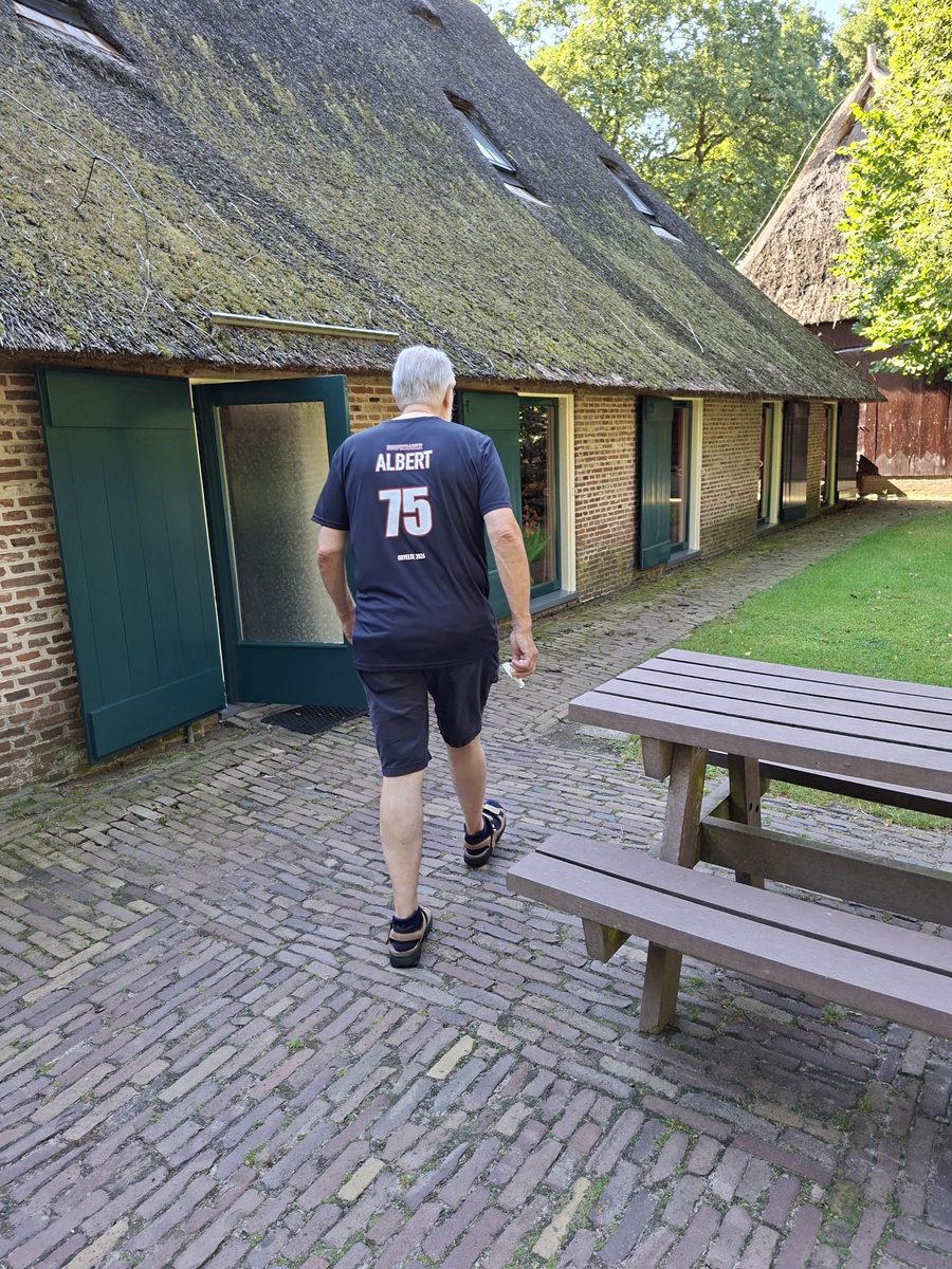 Albert loopt van de camera weg naar de rieten schuur, op de rug van zijn shirt staat ALBERT en 75