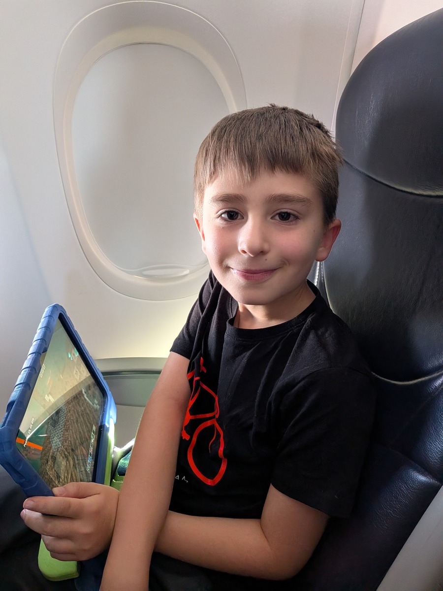 Luca zit lachend in het vliegtuig met een tablet op schoot, het raampje achter hem