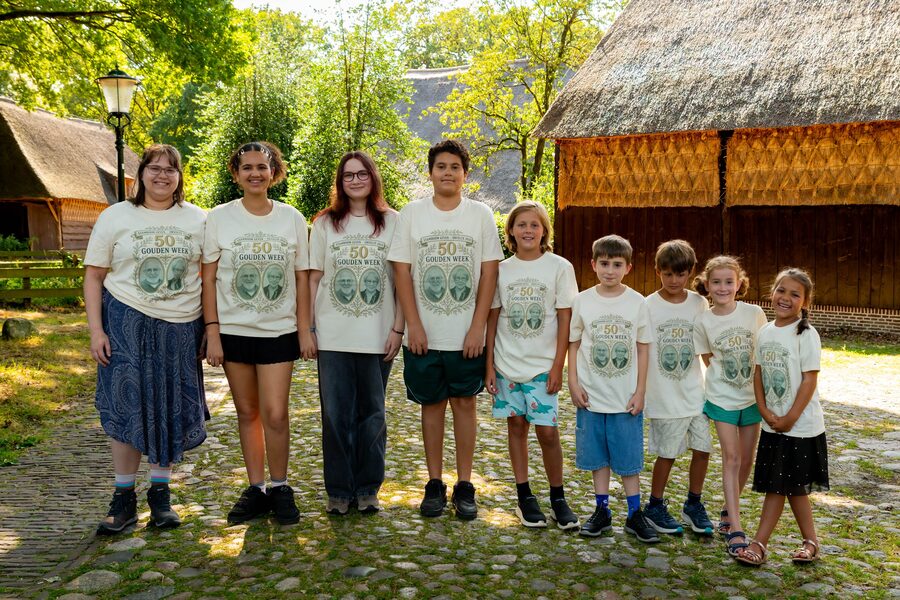 De negen kleinkinderen naast elkaar op een rij in het crèmekleurige Stamboom-shirt, tussen de rieten boerderijen
