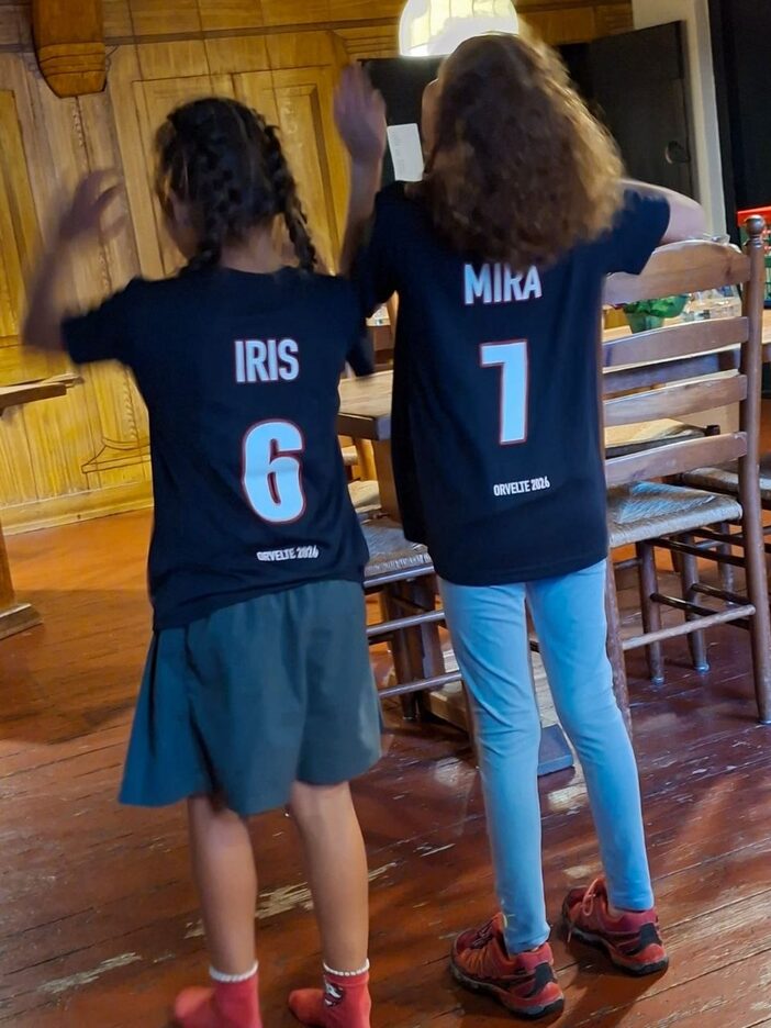 Iris en Mira op de rug gezien in hun donkerblauwe shirt, met IRIS 6 en MIRA 7 en daaronder ORVELTE 2026